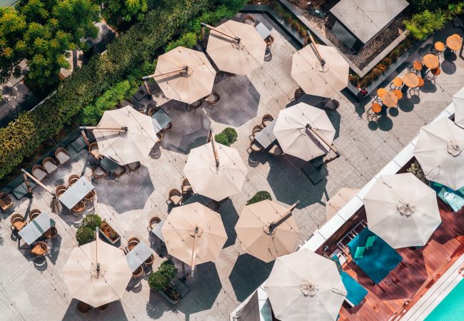 Top view from a drone of an outdoor bar with several open octagonal sunshades making a funny landscape, bordering one part of the terrace a garden, on the other side a lounge area in front of the pool